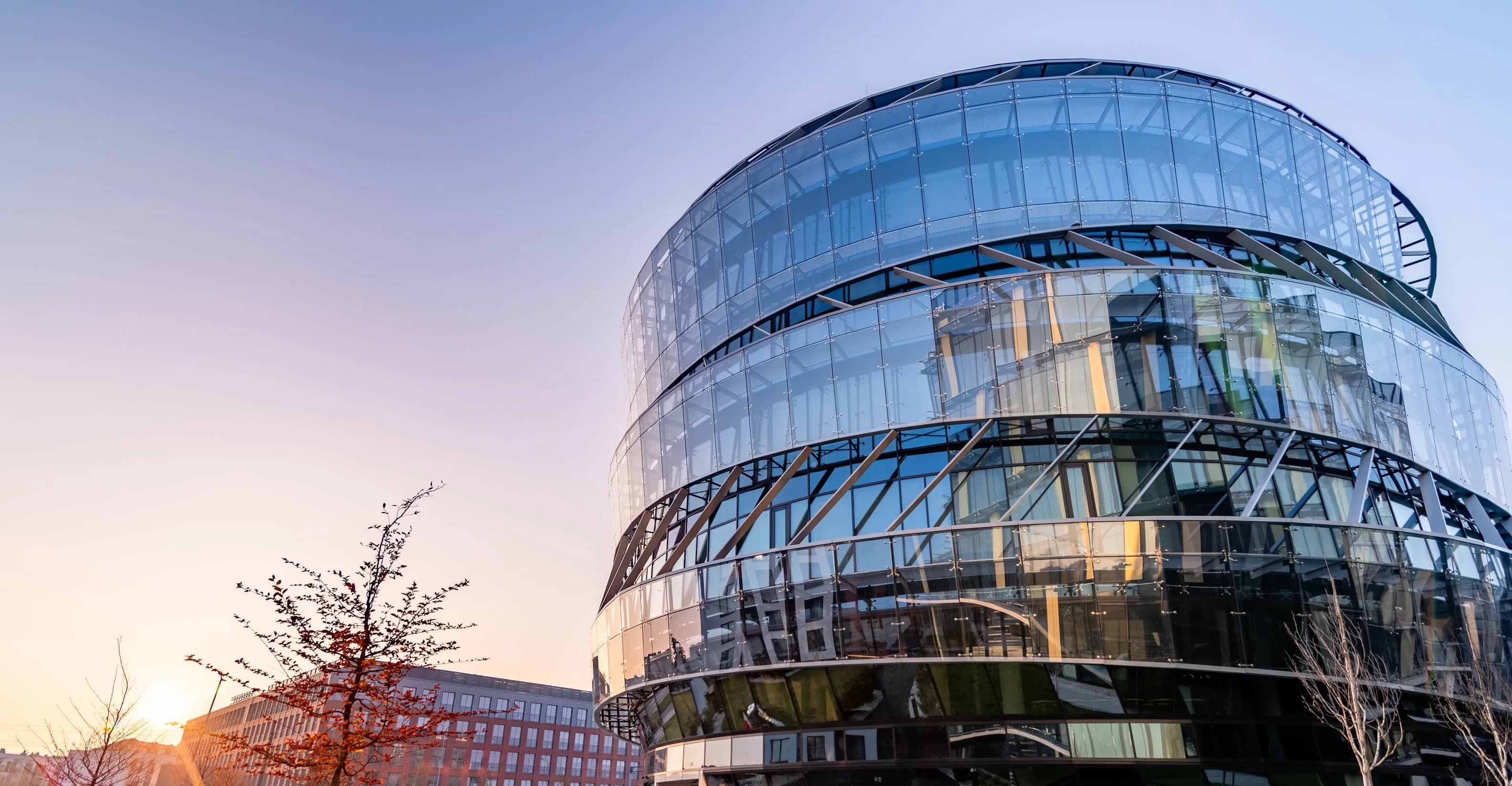 Modern cylindrical glass office building with multiple curved levels reflecting the surrounding cityscape, photographed at sunset with a clear sky and warm light.