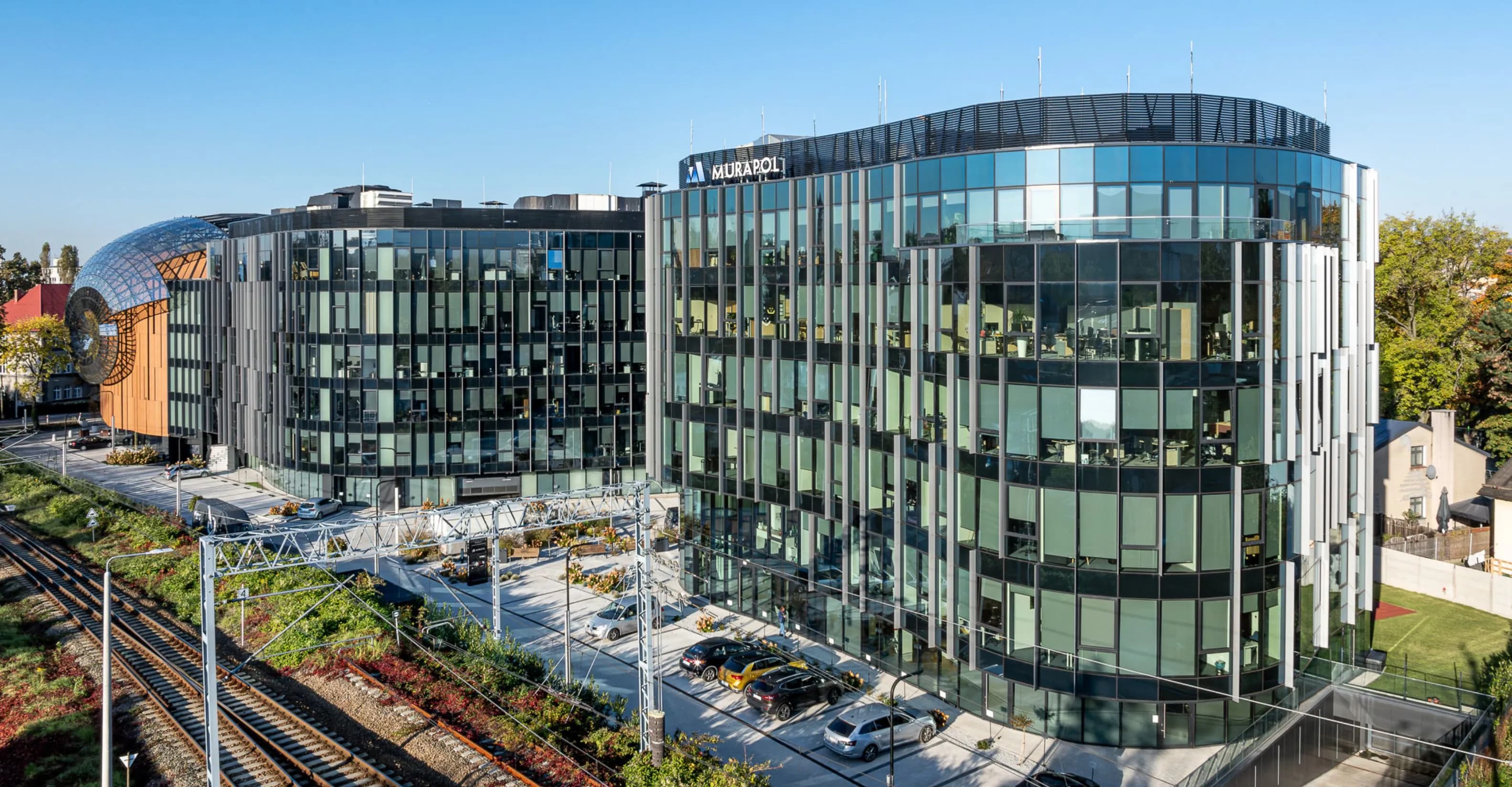 Modern office complex with glass facades reflecting the sky, located next to railway tracks, featuring a distinctive wooden dome structure and surrounding greenery.
